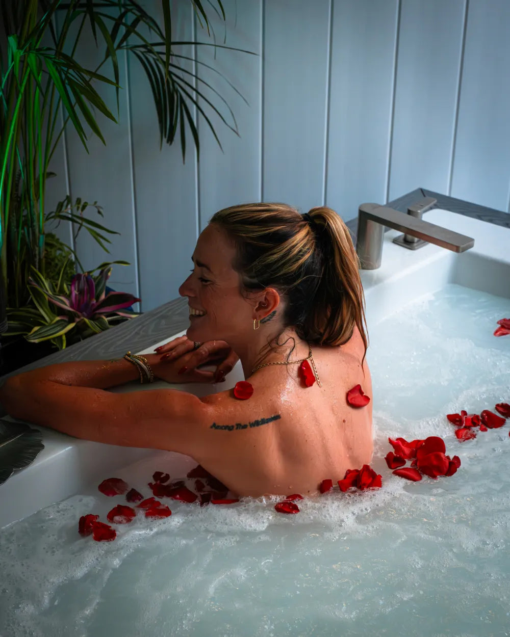 woman relaxing in a bubble bath with rose petals, smiling and leaning on the tub’s edge